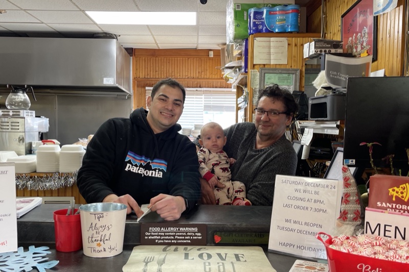 Three generations of the Labrakis family behind the counter at Santoro&rsquo;s Pizza on Christmas Eve, 2022 &mdash; Nicholas, his infant son, and John.